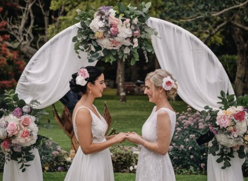 Two brides in white dresses under floral arch in garden